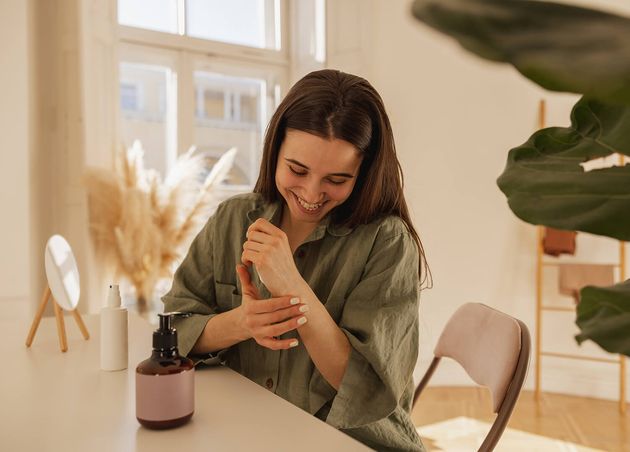 Cheerful Young Caucasian Woman Applies Cream Her Hands while Sitting Table Light Room Brunette Girl with Smooth Skin Wears Shirt Wellness Self Care Concept