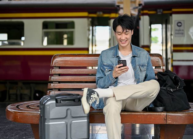 Happy Asian Male Backpacker Using His Phone while Waiting His Train Bench Platform