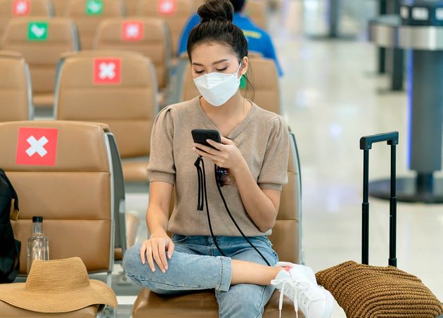 Young Asian Adult Traveller Wearing Face Mask Hand Hold Smartphone Sitting with Social Distancing Wait Flight Schedule Lobby Area Airport Terminal Safety Travel Ideas Concept