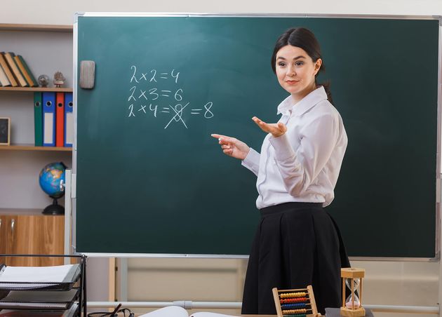 Pleased Holding out Hand Camera Young Female Teacher Standing Front Blackboard Write Classroom