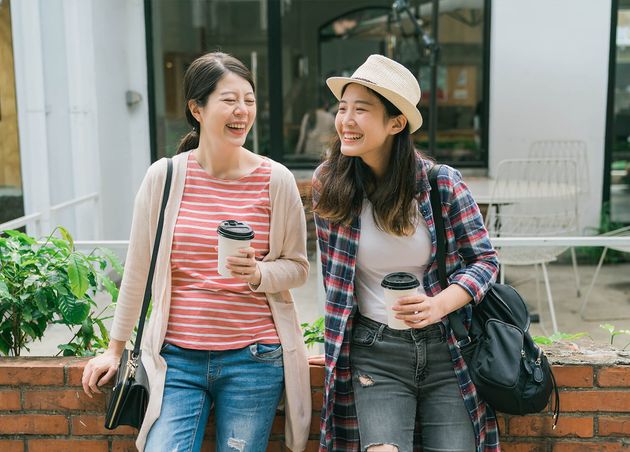 Two Asian Japanese Female Friends Having Drinks Outdoors during Travel Bangkok Thailand Happy Women Tourists Leaning Red Brick Wall Laughing Cheerful Girls Holding Paper Cups Coffee