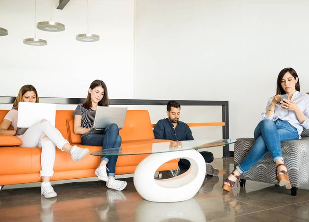 Vier junge lateinamerikanische Menschen sitzen im Bürolounge, verwenden Laptops und Handys und entspannen sich während einer Arbeitspause