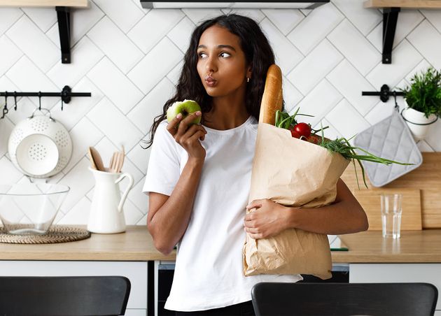 Thoughtful Mulatto Woman Is Holding Package Full with Fresh Vegetables One Hand Bitten Apple Other Modern White Kitchen