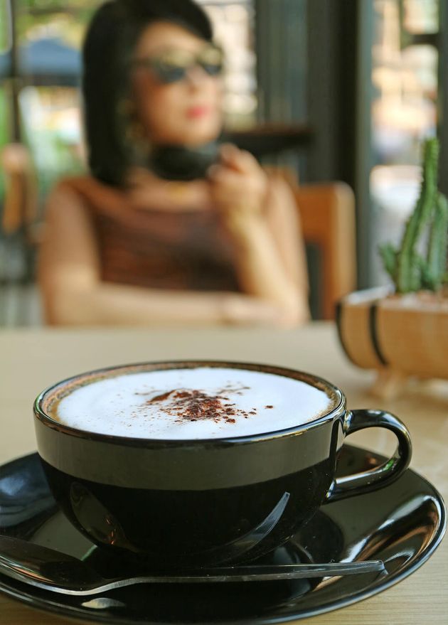 Closeup Cappuccino Black Cup with Blurry Woman Drinking Coffee Backdrop