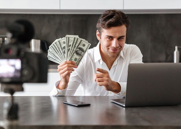 Smiling Young Man Filming His Video Blog Episode about New Tech Devices while Sitting Kitchen Table with Laptop Showing Bunch Money Banknotes