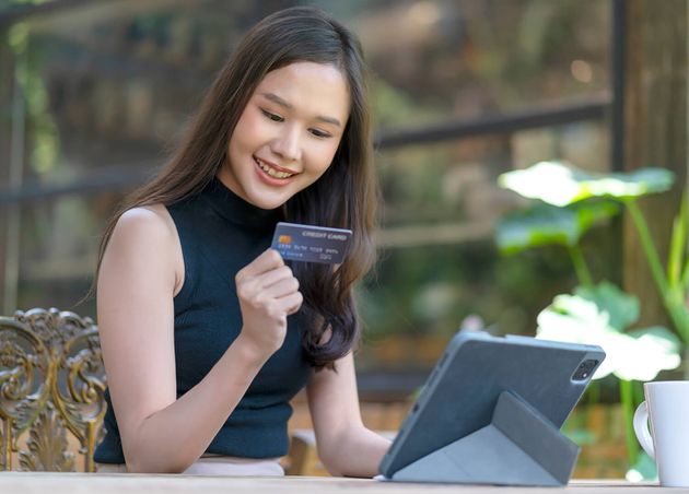 Attraktive asiatische Frau ist glücklich, online mit Laptop einzukaufen, hält Hand mit Kreditkarte, Smartphone, Kaufprodukt mit Hintergrund Café Restaurant, Geschäftsideenkonzept