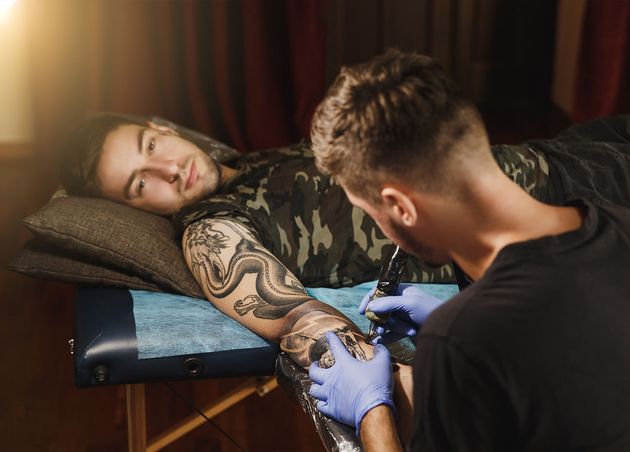 Professional Tattooer Artist Doing Tattoo Arm Young Man by Machine with Black Ink